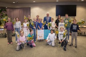 de kinderraadsleden en de burgemeester. De burgemeester staat in het midden. De kinderen staan of hurken naast en voor hem.
